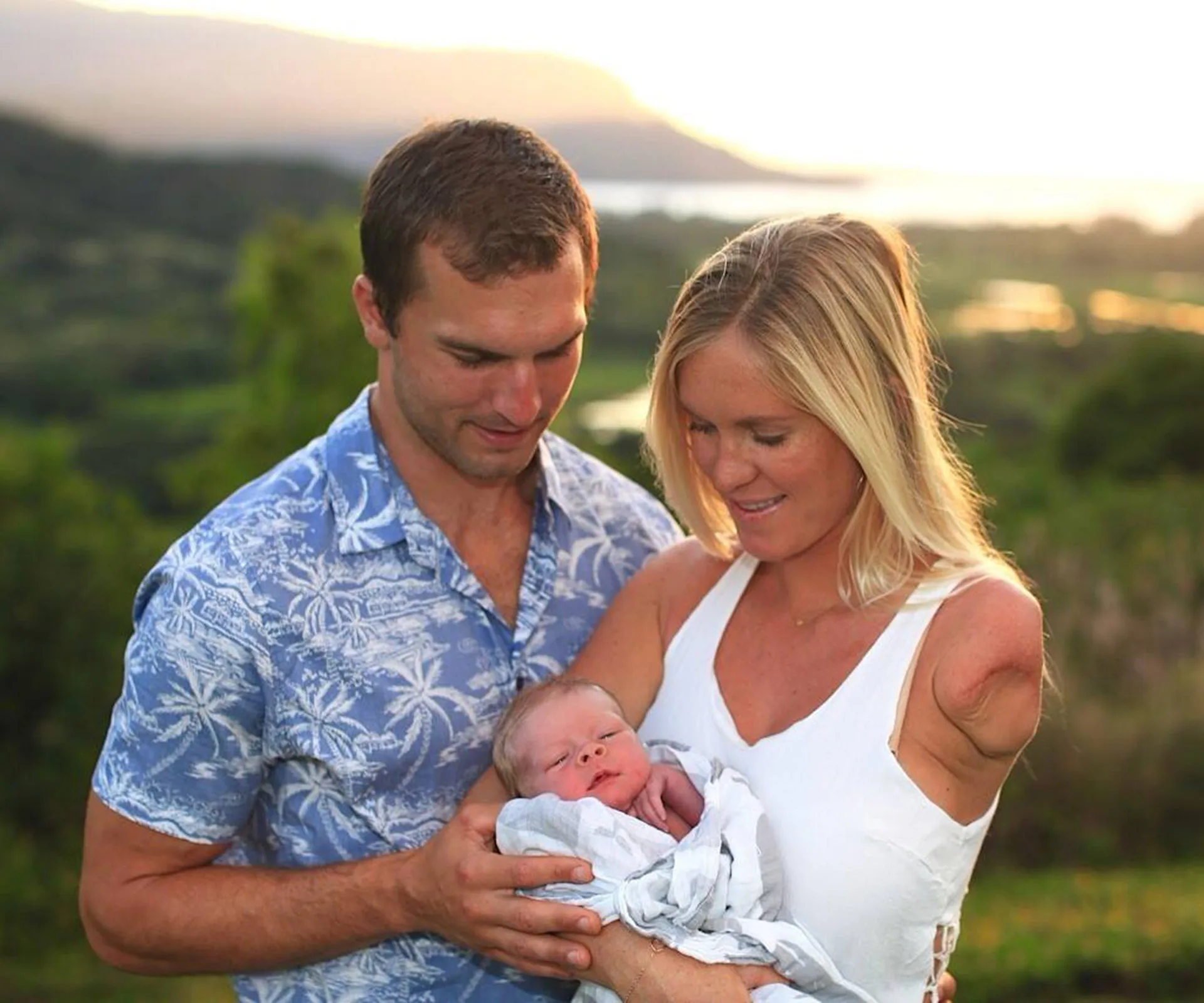 Couple smiling, holding newborn wrapped in a blanket outdoors during sunset.