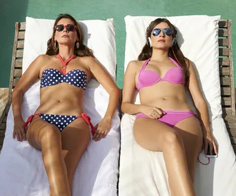 Two women sunbathing near a pool, wearing bikinis and sunglasses. One has headphones and holds a phone.