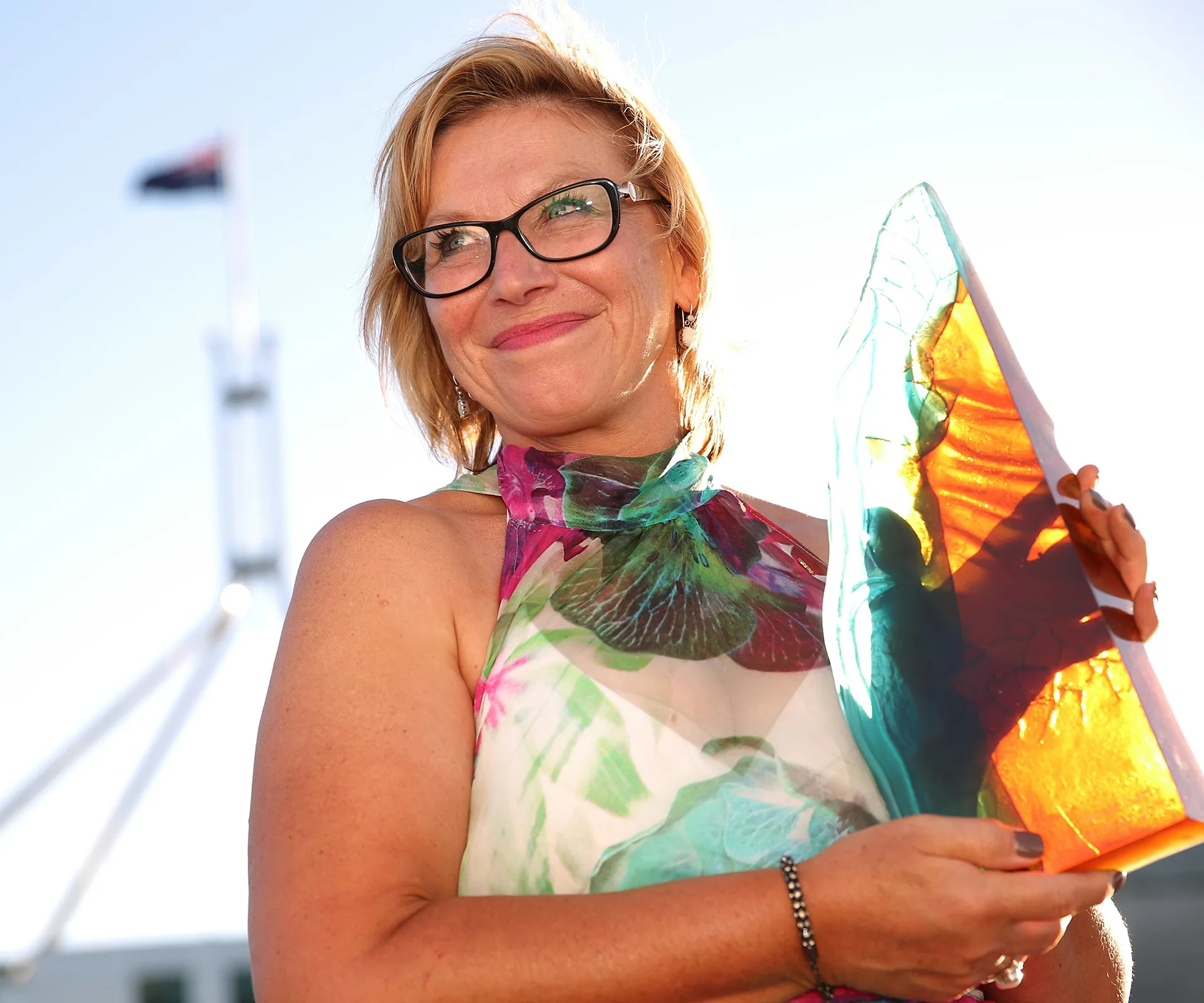 Woman in floral dress holds colorful glass award, with flag and parliament building in the background.