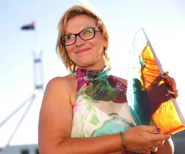 Woman in floral dress holds colorful glass award, with flag and parliament building in the background.