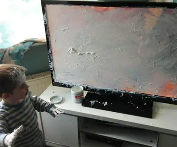 Child covers TV screen with white paint, messy hands and face, container of paint on table.