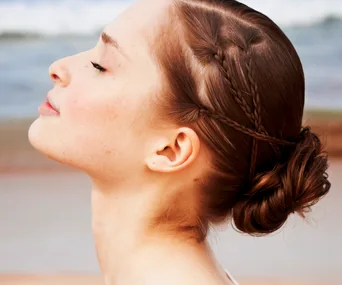 Woman with braided hair bun and closed eyes, profile view against a blurred sandy beach background.
