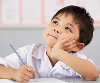 A young boy in a school uniform, holding a pencil, looks thoughtful while writing in a notebook.