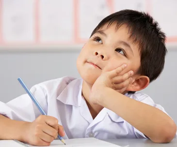 A young boy in a school uniform, holding a pencil, looks thoughtful while writing in a notebook.
