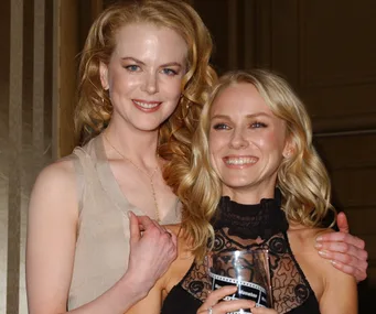 Two women smiling at an event, one holding a glass award, standing close together in evening attire.