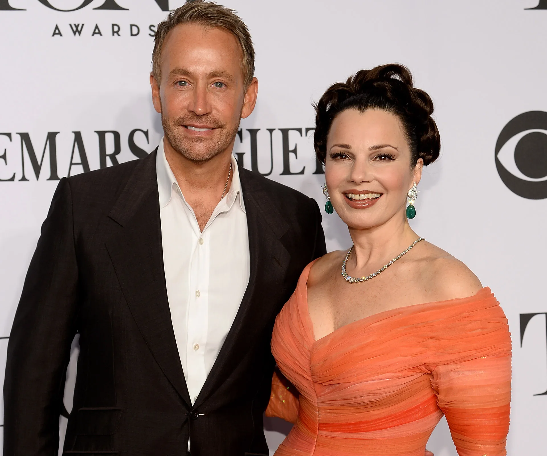 "Two people posing together on a red carpet event; one in a suit, the other in an orange gown with elegant jewelry."