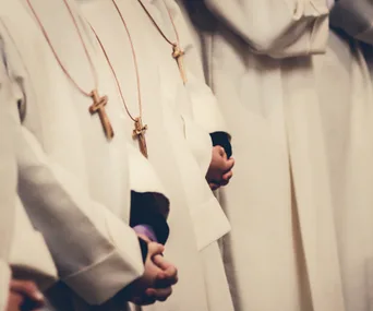 Young people in white robes, wearing cross necklaces, standing in a row with hands clasped.