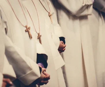 Young people in white robes, wearing cross necklaces, standing in a row with hands clasped.