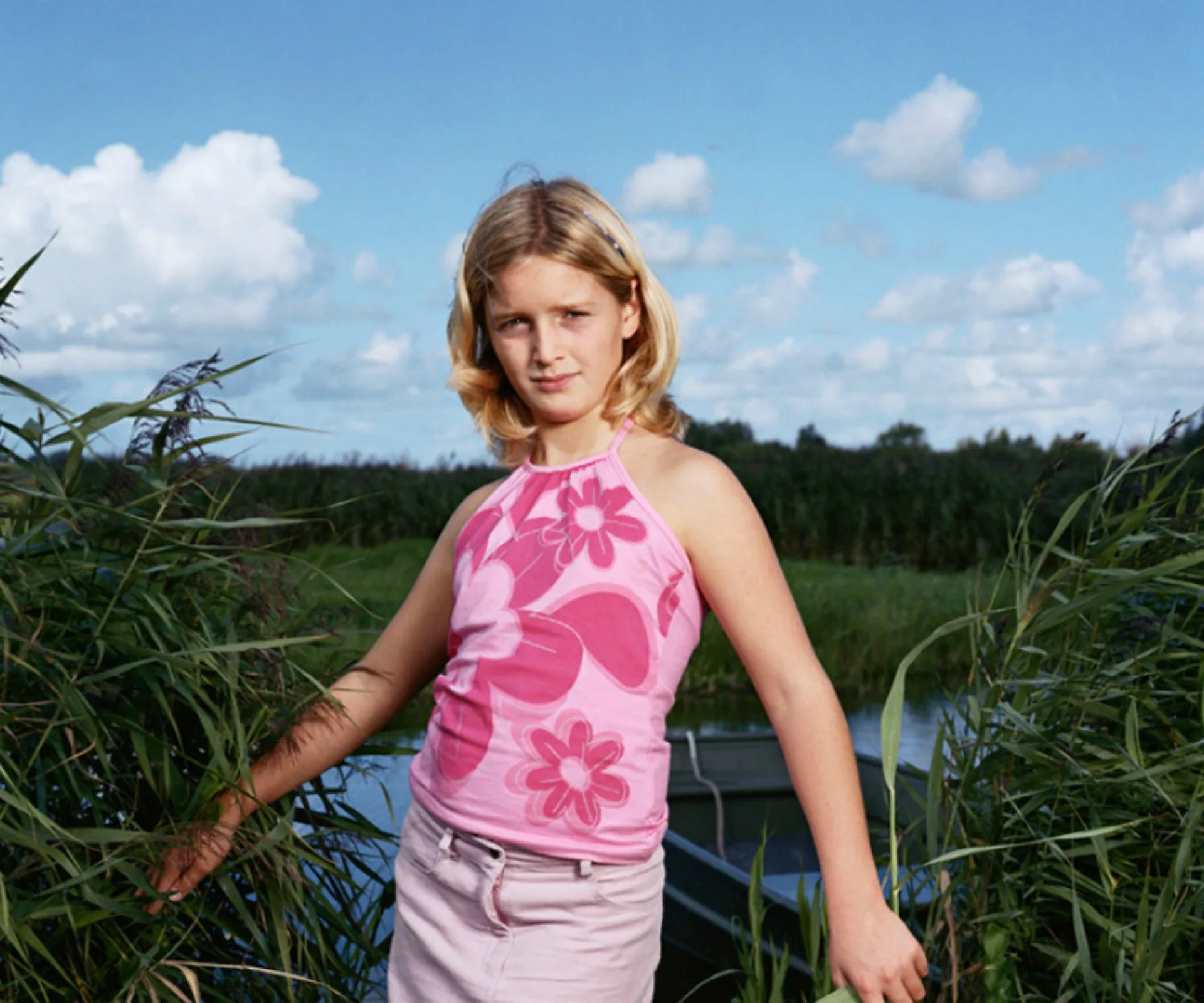 Blonde child in a pink floral top stands amid greenery with a blue sky and clouds in the background.