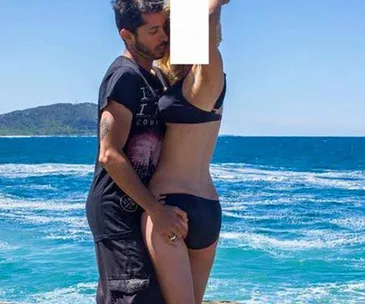 Couple in swimwear embracing on a beach with the ocean and a hill in the background.