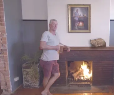 Man with beer leaning on a fireplace, portrait above mimics pose, cat figurine on mantle.