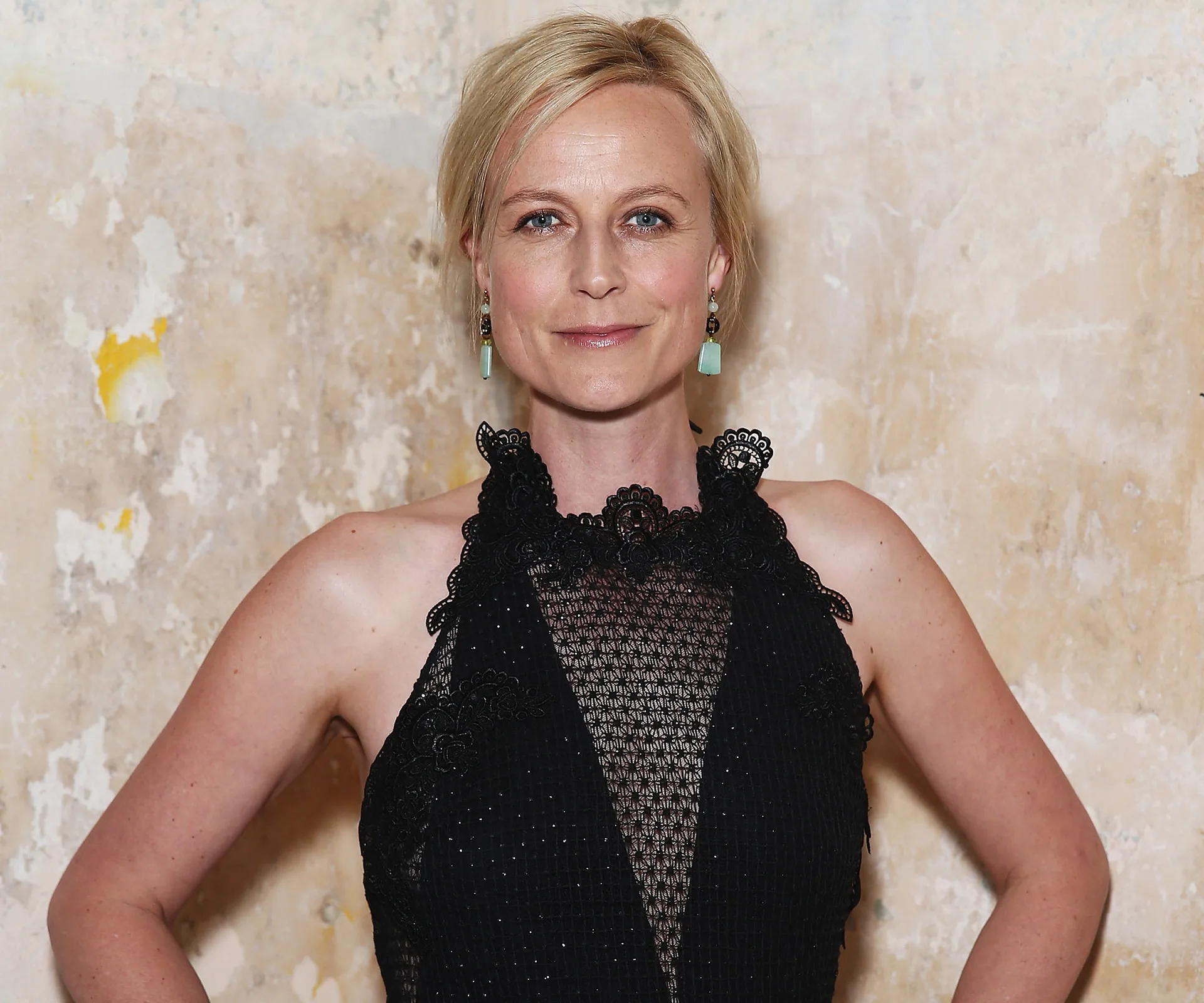 A person wearing a black dress stands against a textured wall.