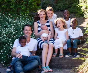 A family of eight sits together outdoors, smiling, with greenery and flowers in the background.