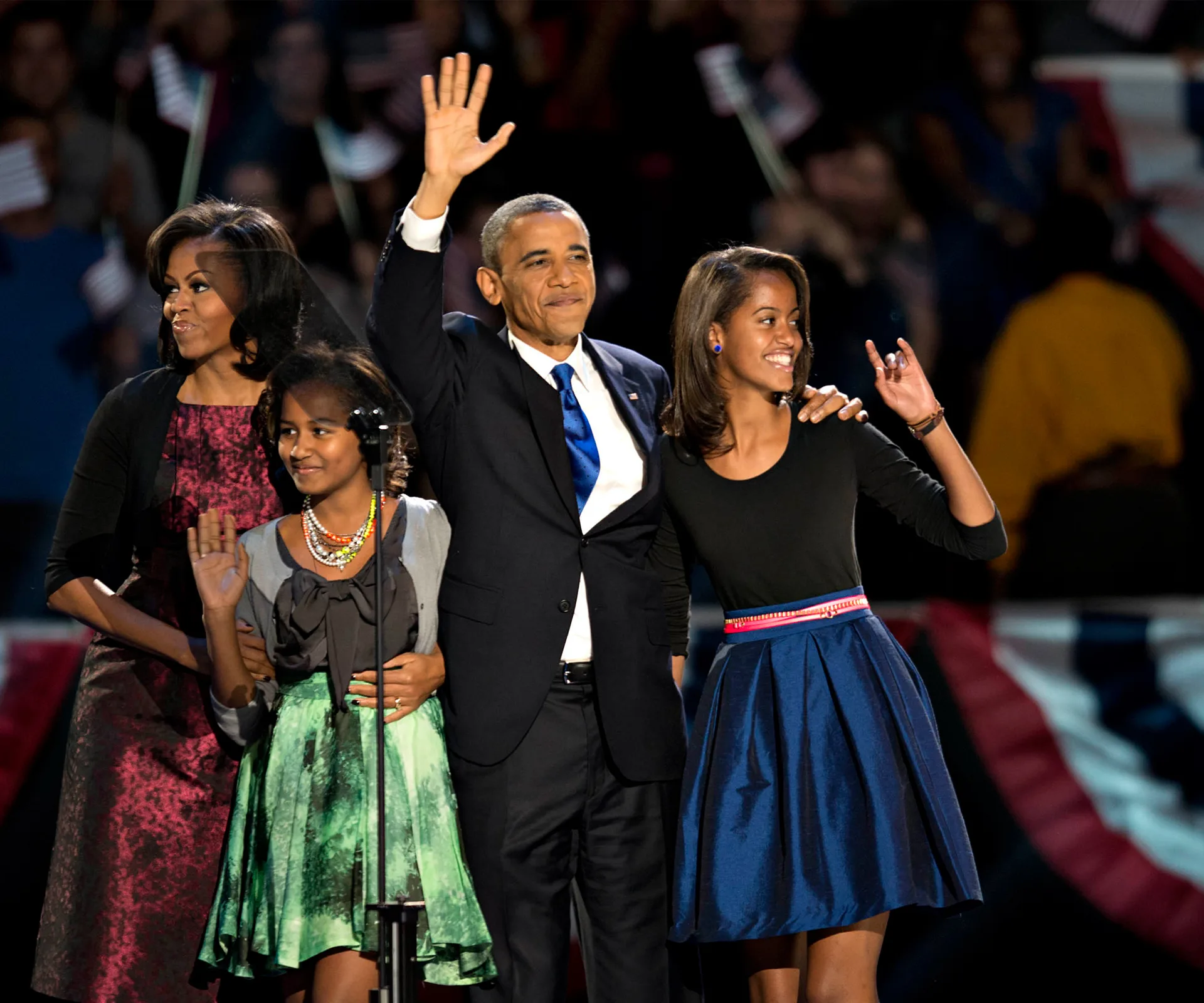A family stands together on stage, smiling and waving at a public event, with patriotic decorations in the background.