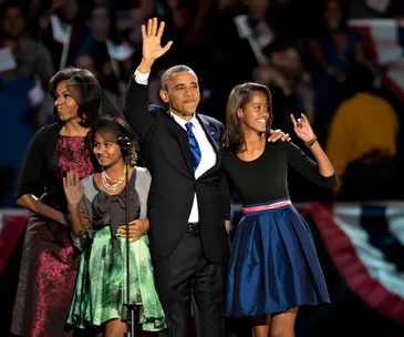 A family stands together on stage, smiling and waving at a public event, with patriotic decorations in the background.