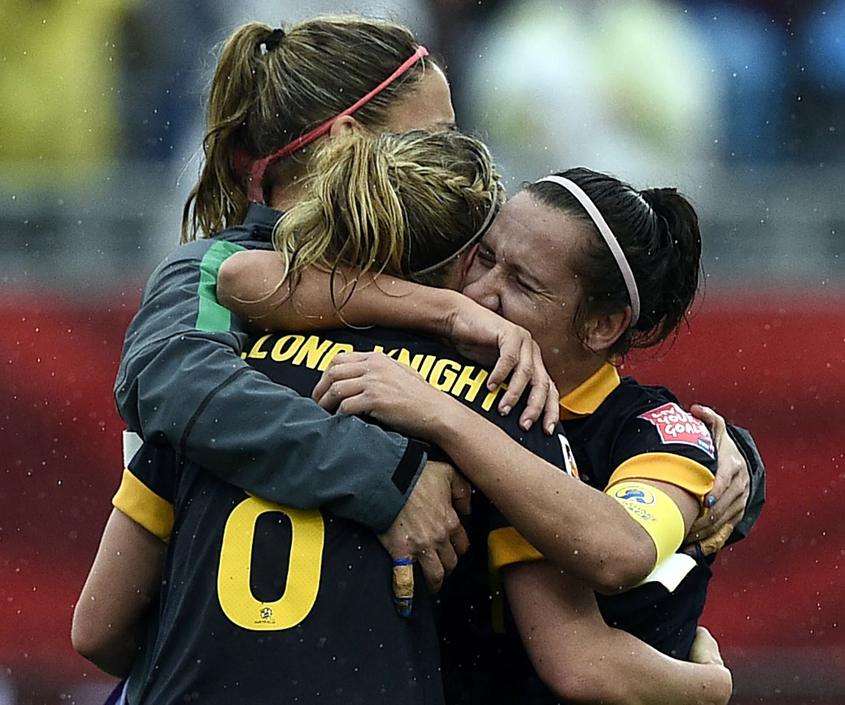 Three soccer players in uniform embrace emotionally on a rainy field, celebrating a victory.