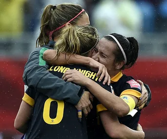 Three soccer players in uniform embrace emotionally on a rainy field, celebrating a victory.