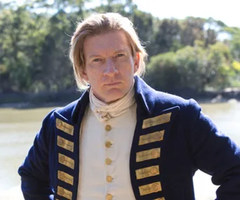 A character in a blue historical military uniform stands outdoors by a river, with trees in the background.