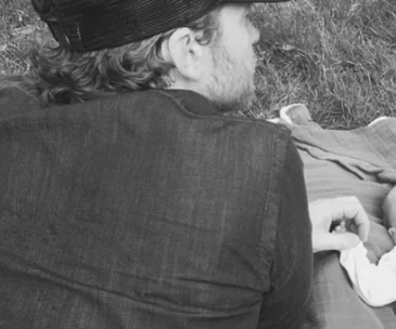 A person lying on grass next to a wrapped baby, holding its hand in an intimate moment.