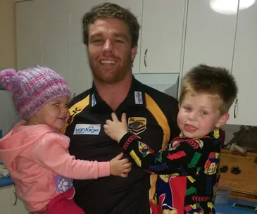Rugby player holding two smiling children in a kitchen, one wearing a colorful pajama and the other a pink outfit with a beanie.