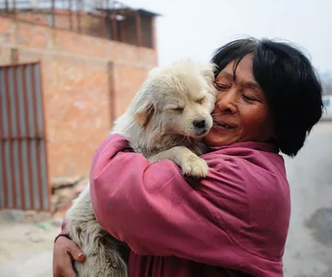 A woman in a pink coat joyfully hugs a small white dog outdoors.