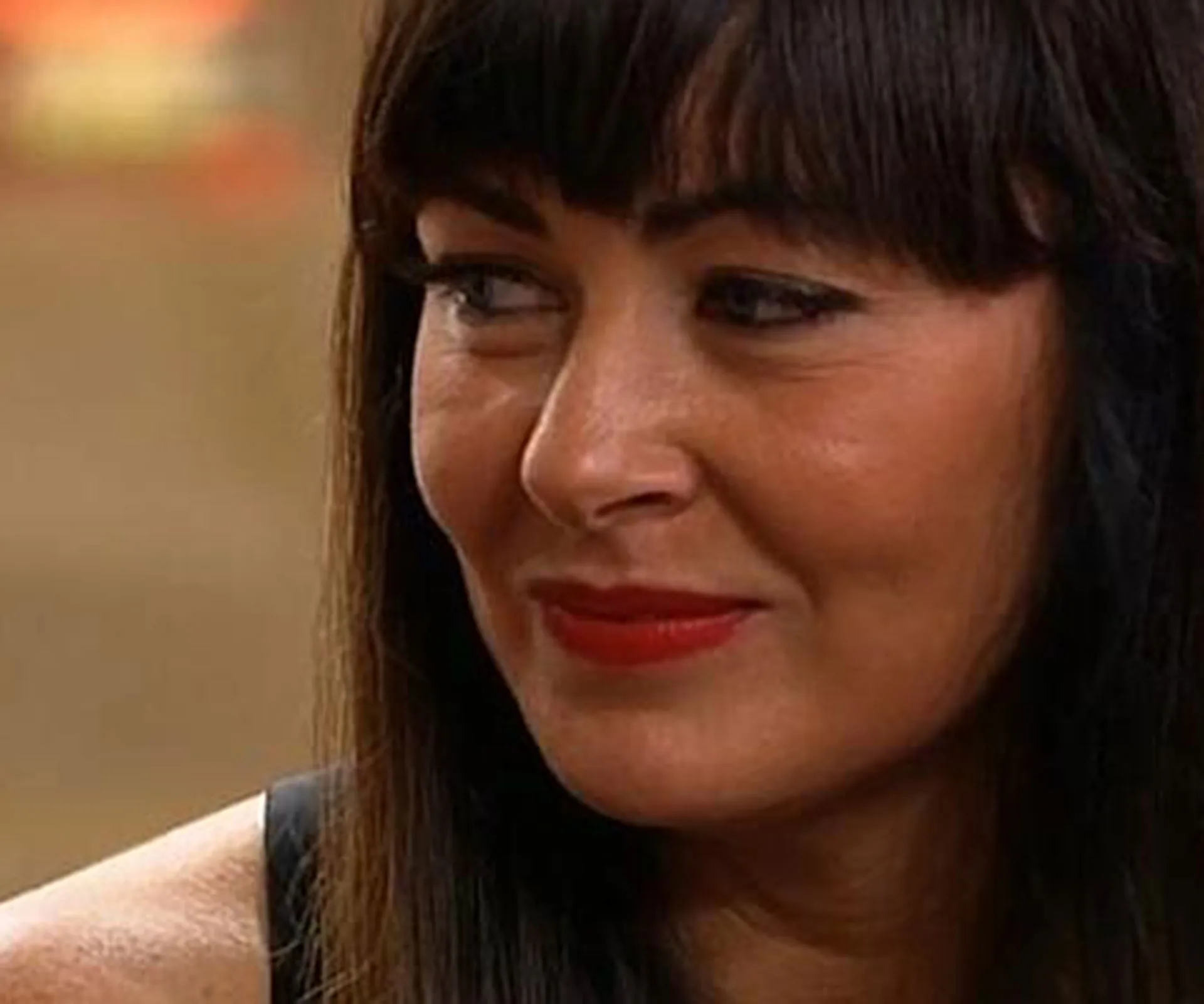 Person with dark hair and red lipstick, smiling slightly, looks to the side on a blurred background.