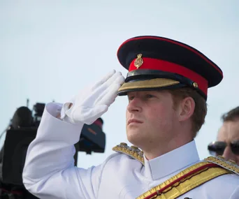 Royal figure in military uniform saluting, wearing a red and black peaked cap, with focused expression.