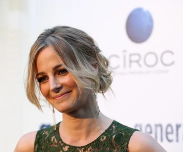 A smiling woman with blonde hair styled in an updo, wearing a green lace dress and long earrings, at an event backdrop.