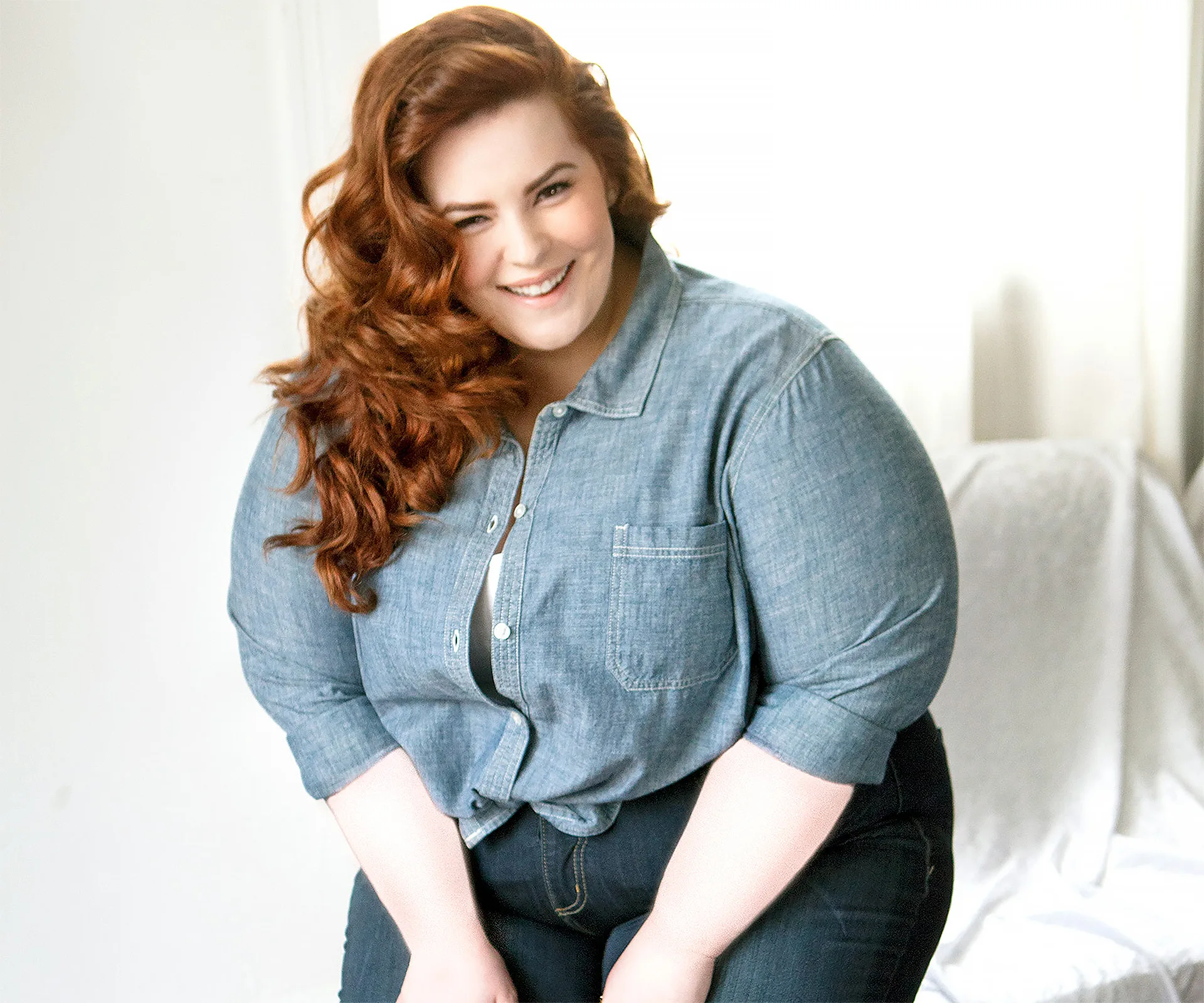 Red-haired woman smiling in a denim shirt, leaning slightly forward against a light background.