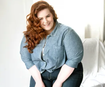 Red-haired woman smiling in a denim shirt, leaning slightly forward against a light background.