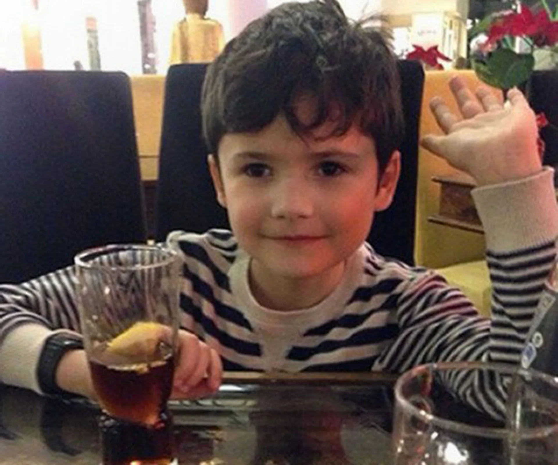 Young boy sitting at a table, wearing a striped shirt, raising his hand, with a drink in front of him.