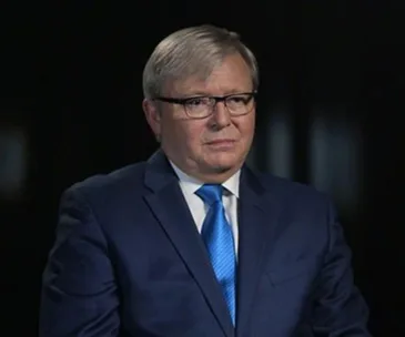 An older man in a suit and glasses, with a serious expression, against a dark background.