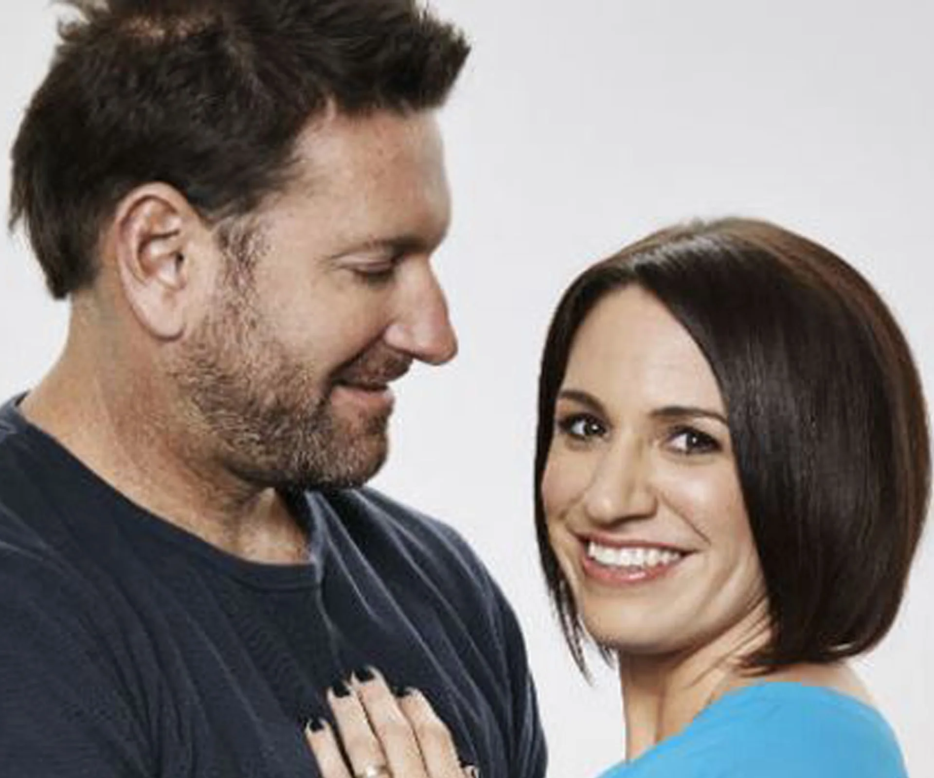 A smiling couple standing close, the man looking at the woman who faces the camera, both appearing cheerful.