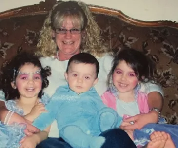 A woman with glasses sitting on a couch with three smiling children on her lap, wearing colorful outfits.