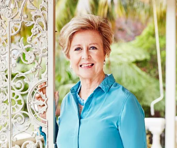 A woman in a teal blouse stands smiling by an ornate white gate with lush greenery in the background.