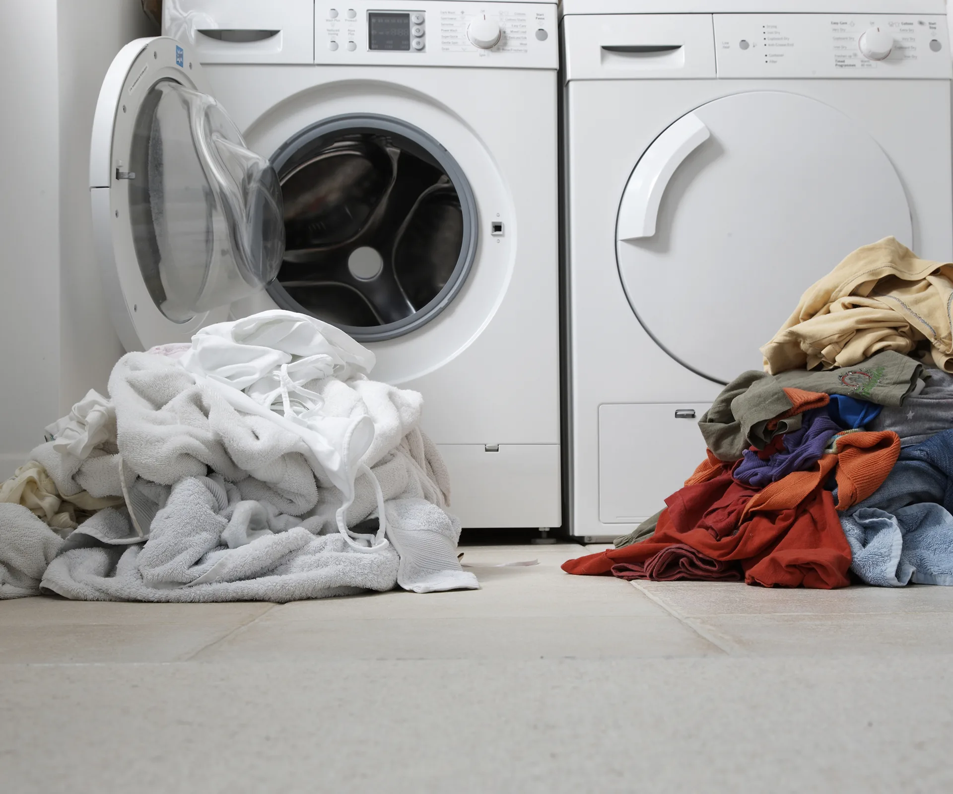 Washing machine and dryer with piles of laundry on the floor, some white and some colored clothes.