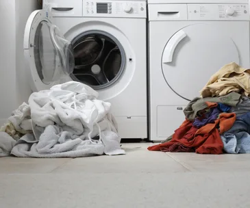Washing machine and dryer with piles of laundry on the floor, some white and some colored clothes.