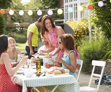 Group of friends enjoying a backyard barbecue with colorful lanterns and a table set with food and drinks.