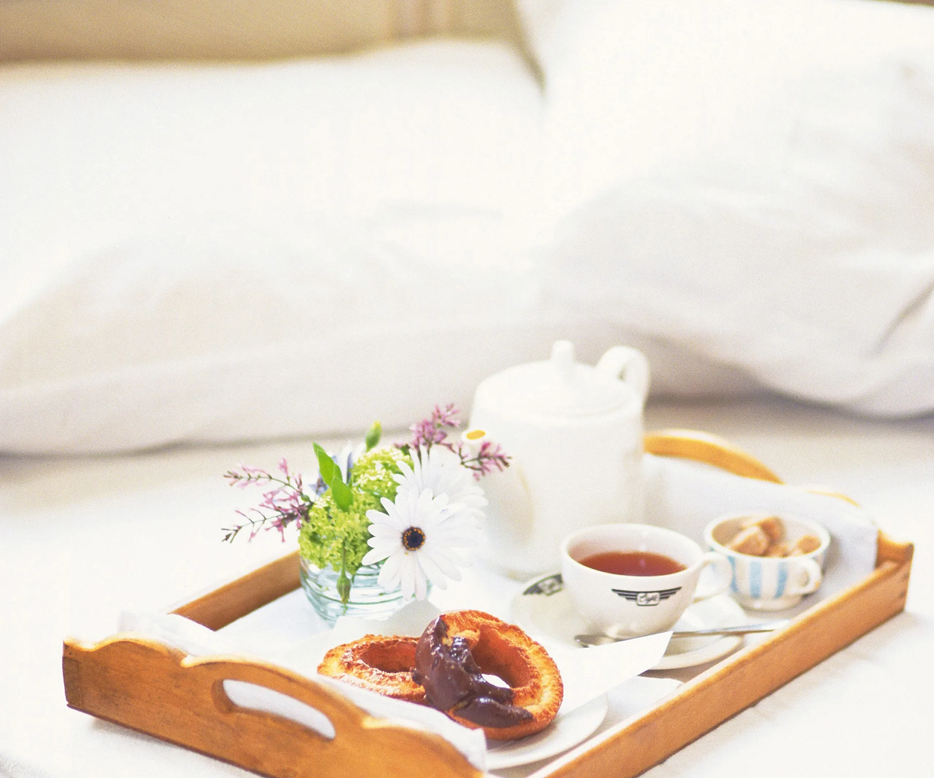 Breakfast tray with tea, pastries, flowers on bed for a cozy morning treat.