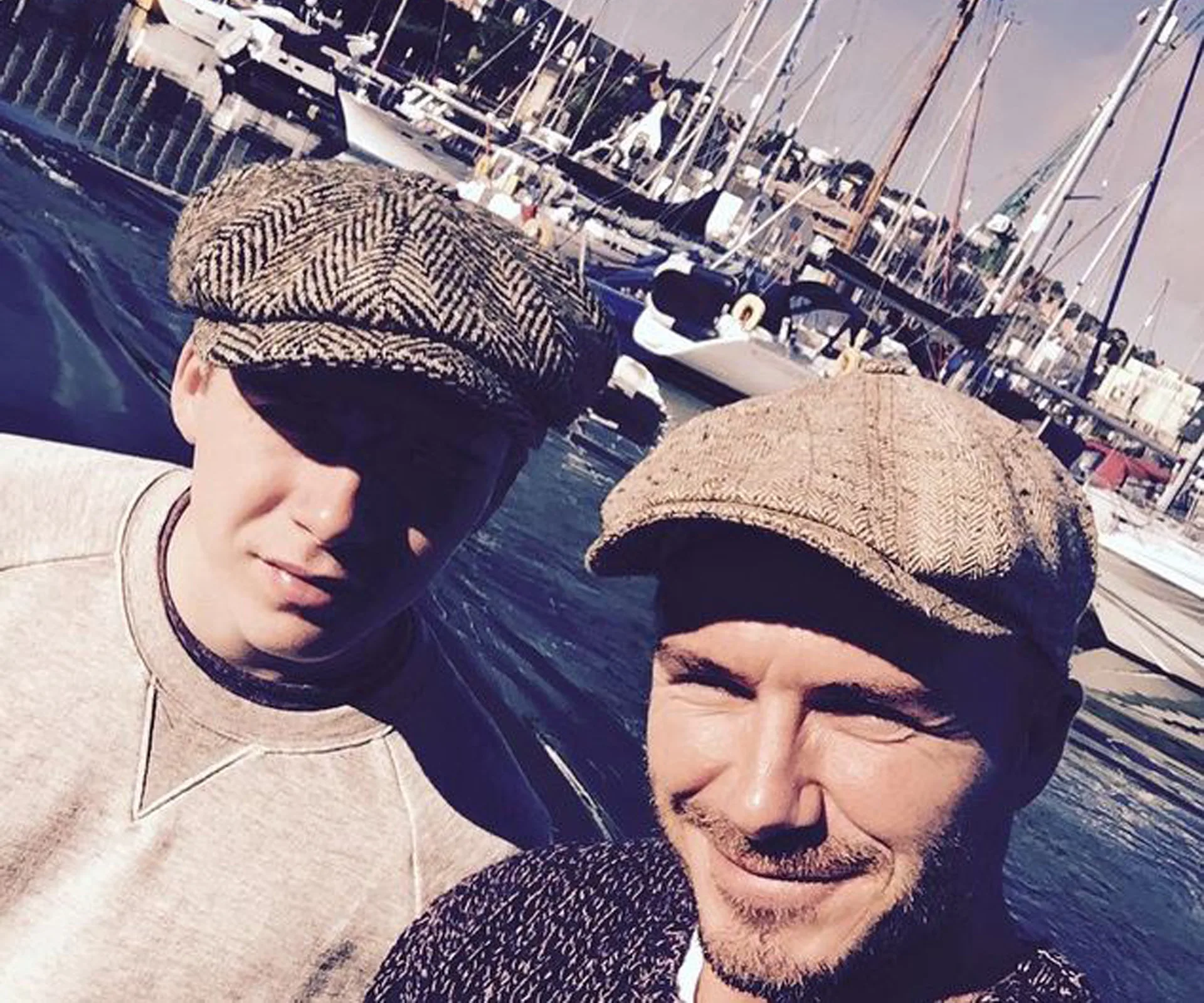Two people wearing caps on a sunny day at a marina with boats in the background.
