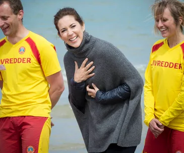 Royal figure in a gray sweater smiles with lifeguards at the beach.