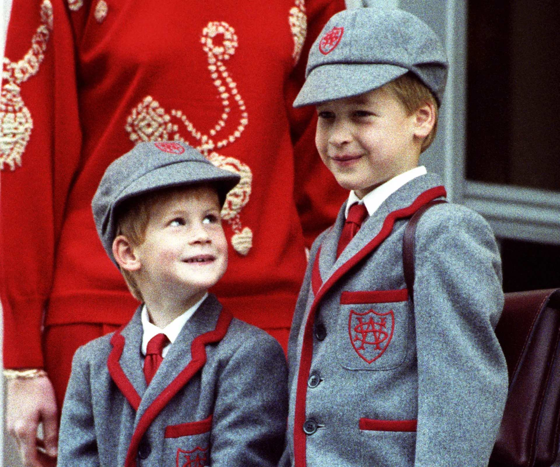 Two young boys in matching gray school uniforms with red trim and caps, standing together, smiling.