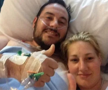 Hospitalized man with oxygen and IVs gives thumbs up, lying in bed beside a woman who also gives a thumbs up.