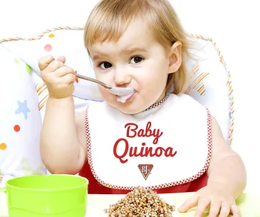 Baby in high chair wearing "Baby Quinoa" bib, eating with spoon, next to pile of quinoa.