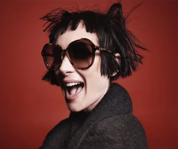 A person with short, spiky black hair and large sunglasses laughs against a red background.