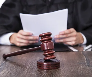 A gavel on a wooden table with a judge in robes reading a document in the background.