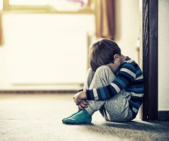 A young boy in striped pajamas sits curled up, leaning against a wall, looking down with a somber expression.