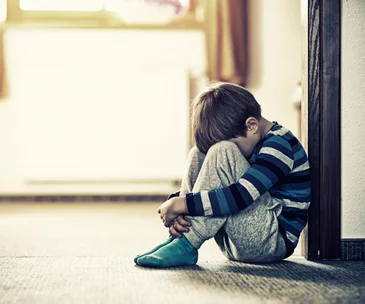 A young boy in striped pajamas sits curled up, leaning against a wall, looking down with a somber expression.