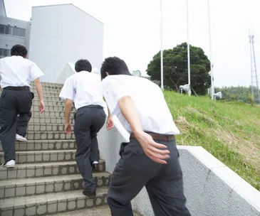Students in uniforms walking up outdoor concrete steps beside a grassy area and flagpoles.