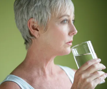 Elderly woman drinking from a glass of water with a neutral expression, against a green background.